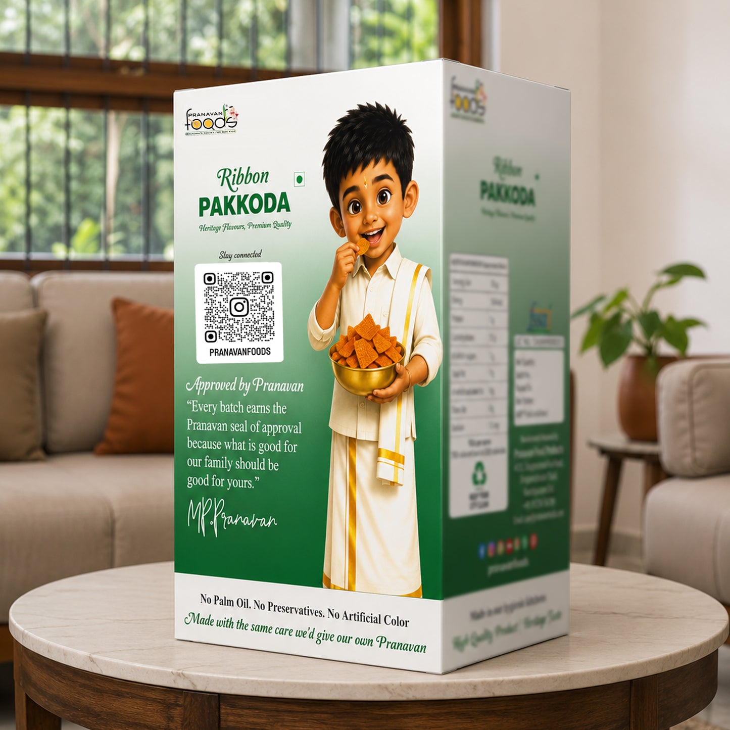 A boxed Pranavan food product labeled 'Ribbon PAKKODA' stands on a round table in a living room setting. The package is predominantly white and green with printed branding, a QR code, and an illustrated child holding a bowl of golden fried snacks.