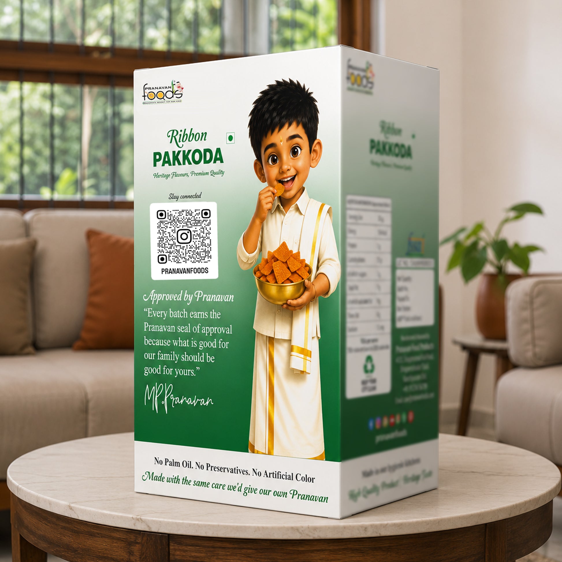 A boxed Pranavan food product labeled 'Ribbon PAKKODA' stands on a round table in a living room setting. The package is predominantly white and green with printed branding, a QR code, and an illustrated child holding a bowl of golden fried snacks.