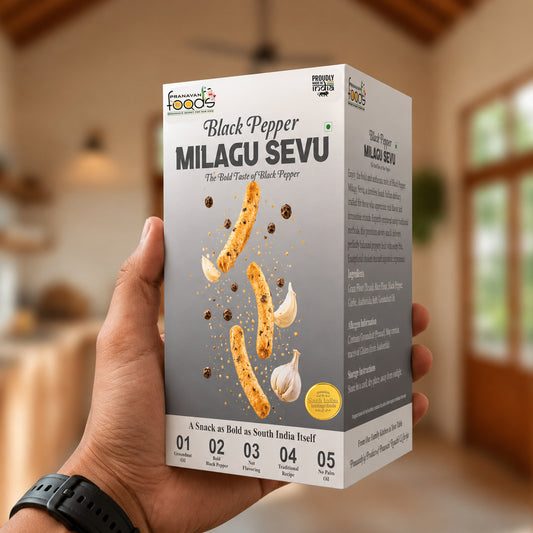 A hand holds a box of Pranavan Foods Black Pepper Milagu Sevu in a warm, blurred indoor setting. The packaging is white and gray with illustrations of sevu, garlic, and black peppercorns.