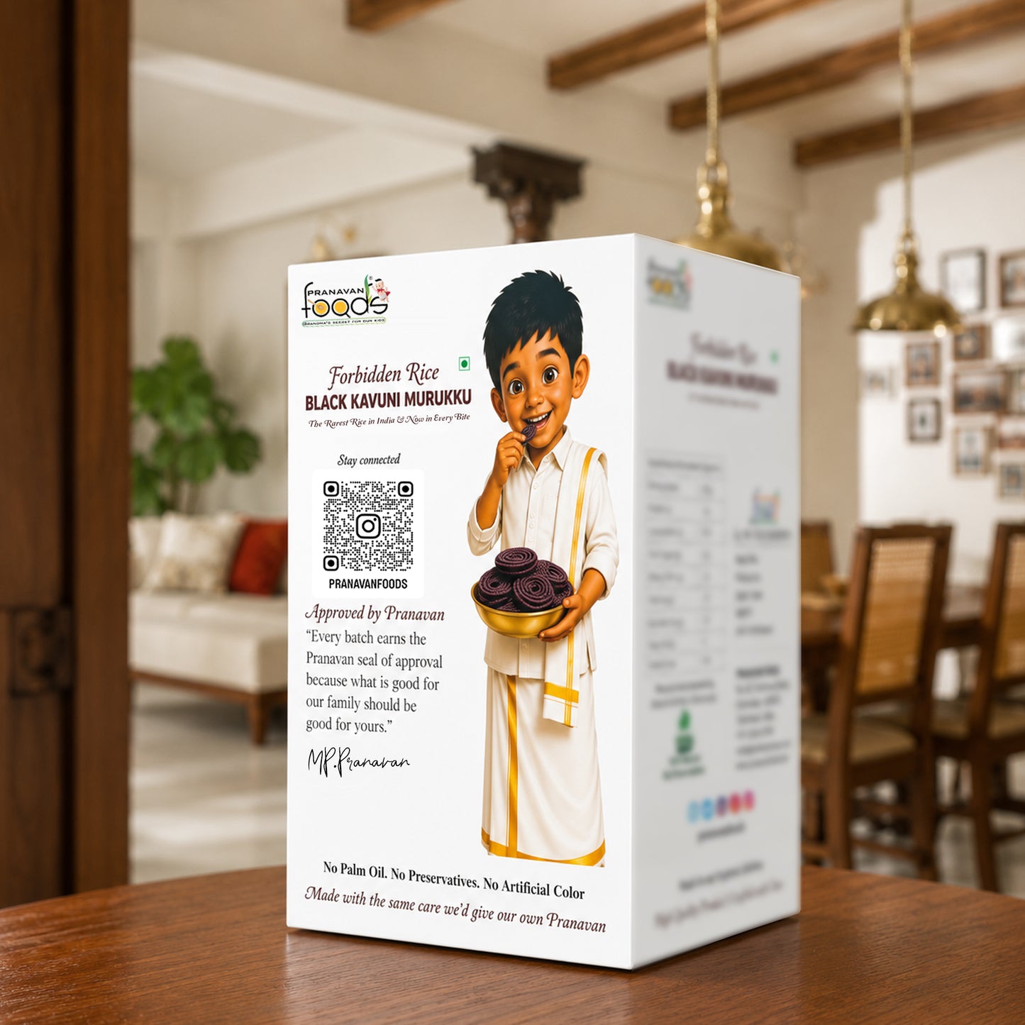 A white product box for Forbidden Rice Black Kavuni Murukku stands on a wooden table in a warm, blurred dining room setting. The box features a cartoon illustration of a boy holding a bowl of dark round snacks, with product details, a QR code, and the Pranavan approval signature printed on the front.