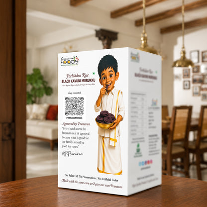 A white product box for Forbidden Rice Black Kavuni Murukku stands on a wooden table in a warm, blurred dining room setting. The box features a cartoon illustration of a boy holding a bowl of dark round snacks, with product details, a QR code, and the Pranavan approval signature printed on the front.