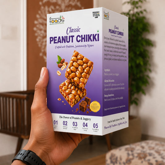 A hand holds a box of Peanut Chikki against an indoor background with a wooden chair and plant. The box is white and purple with images of peanut chikki bars and text detailing the product.