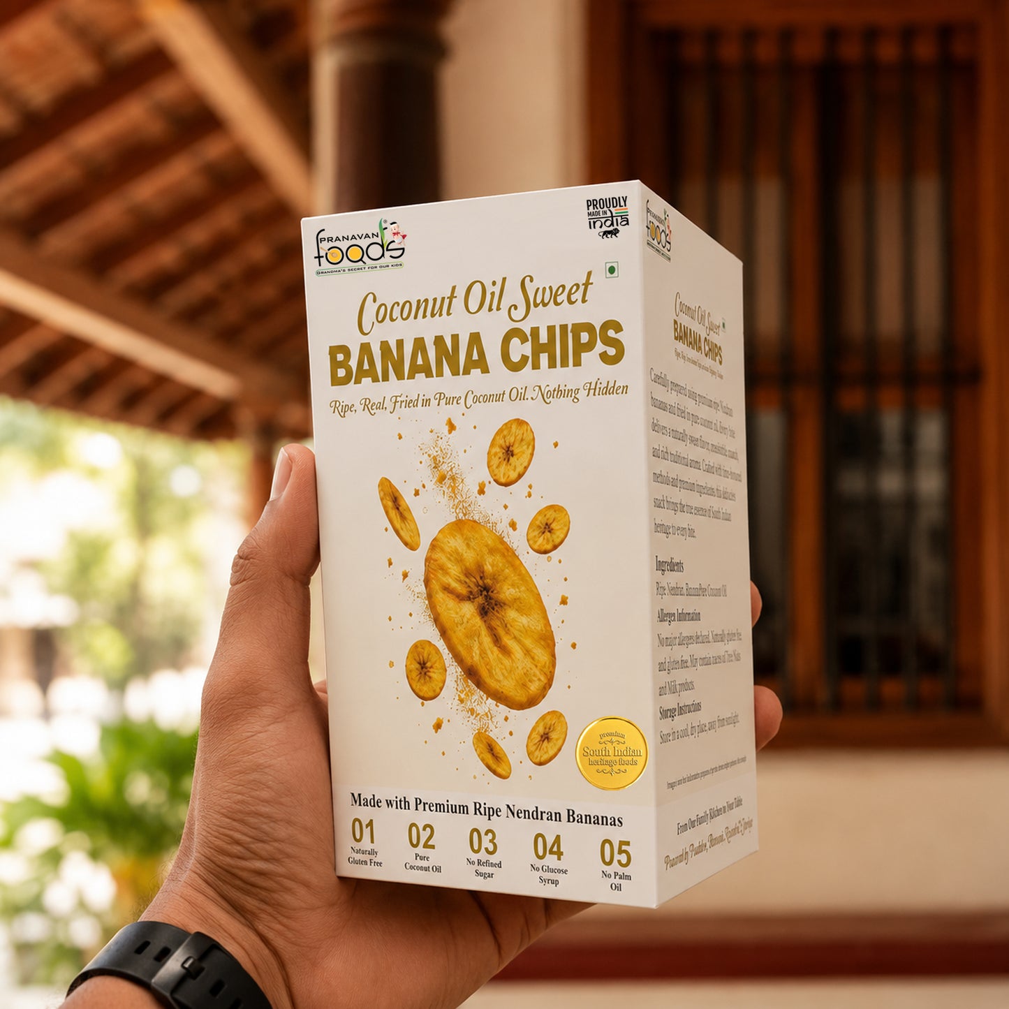 A hand holds a box of Fresh from the Forests Coconut Oil Sweet Banana Chips in front of a tropical wooden structure.