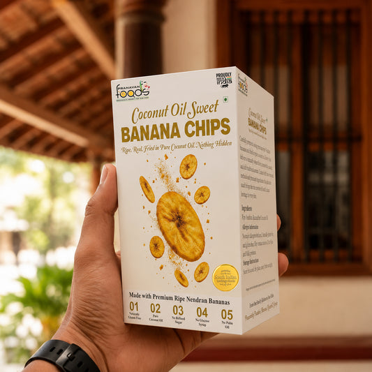 A hand holds a box of Fresh from the Forests Coconut Oil Sweet Banana Chips in front of a tropical wooden structure.