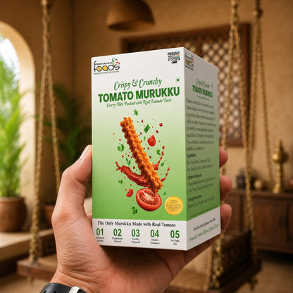 A hand holds a box of Pranavan Foods Tomato Murukku in a warm, rustic indoor setting with hanging decor in the background. The box is white and light green with an illustration of crispy, golden-brown fried fish sticks with tomato and chili splatters.