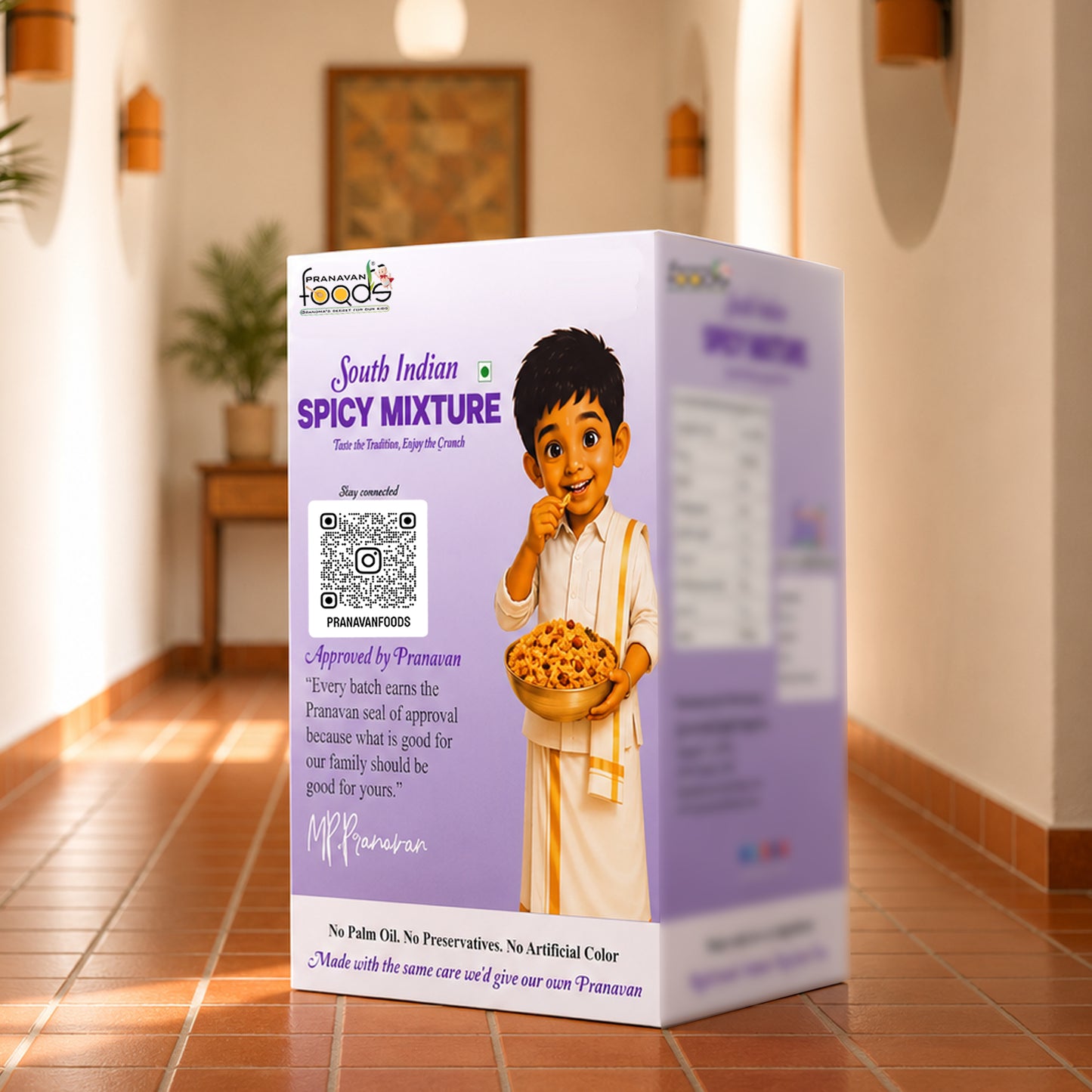 A box of South Indian Spicy Mixture by the 'pranavan foods' is shown on a terracotta tiled floor in a warm, traditional room setting. The packaging is primarily light purple with a cartoon illustration of a boy holding a bowl of food.