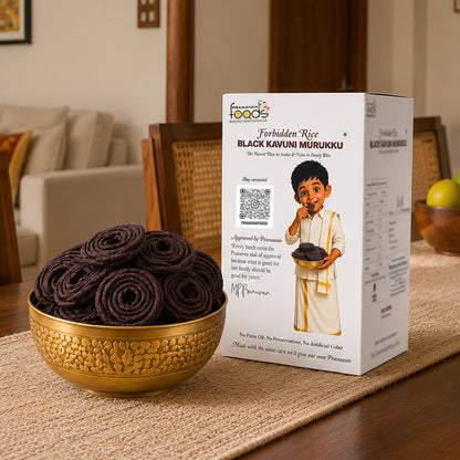 A box of Pranavan Foods Black Kavuni Murukku alongside a gold bowl filled with rolled dark brown/black crispy snacks, set on a wooden table mat in a warm living room environment.