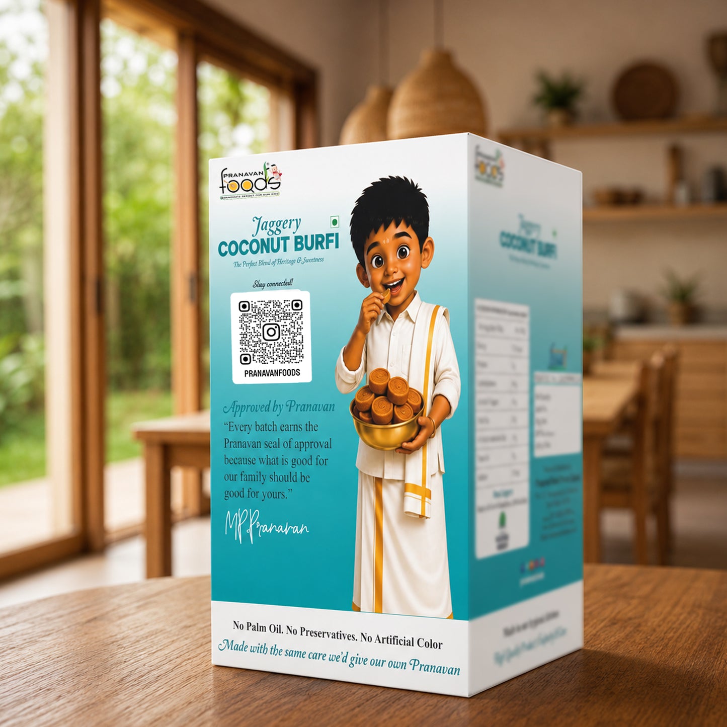 A boxed food product standing on a wooden table in a warm indoor setting. The package is predominantly white and light blue with a cartoon boy holding a bowl of round coconut burfi, and visible text including 'Jaggery Coconut Burfi' and 'Pranavan Foods'.