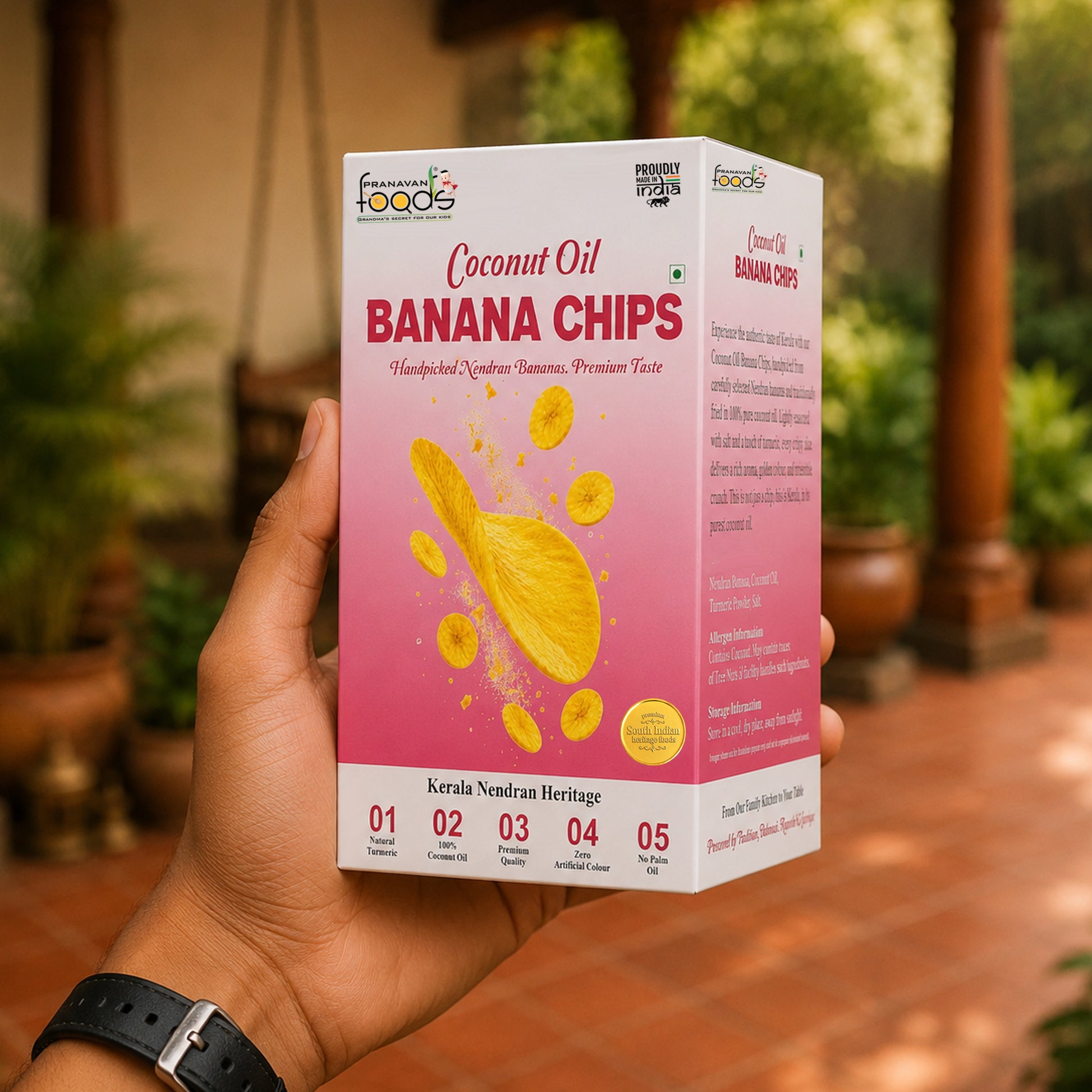 A hand holds a box of Pranavan Foods Banana Chips Coconut Oil in front of a blurred outdoor background. The box is pink and white with yellow banana chip graphics and text indicating Kerala Nendran Heritage.