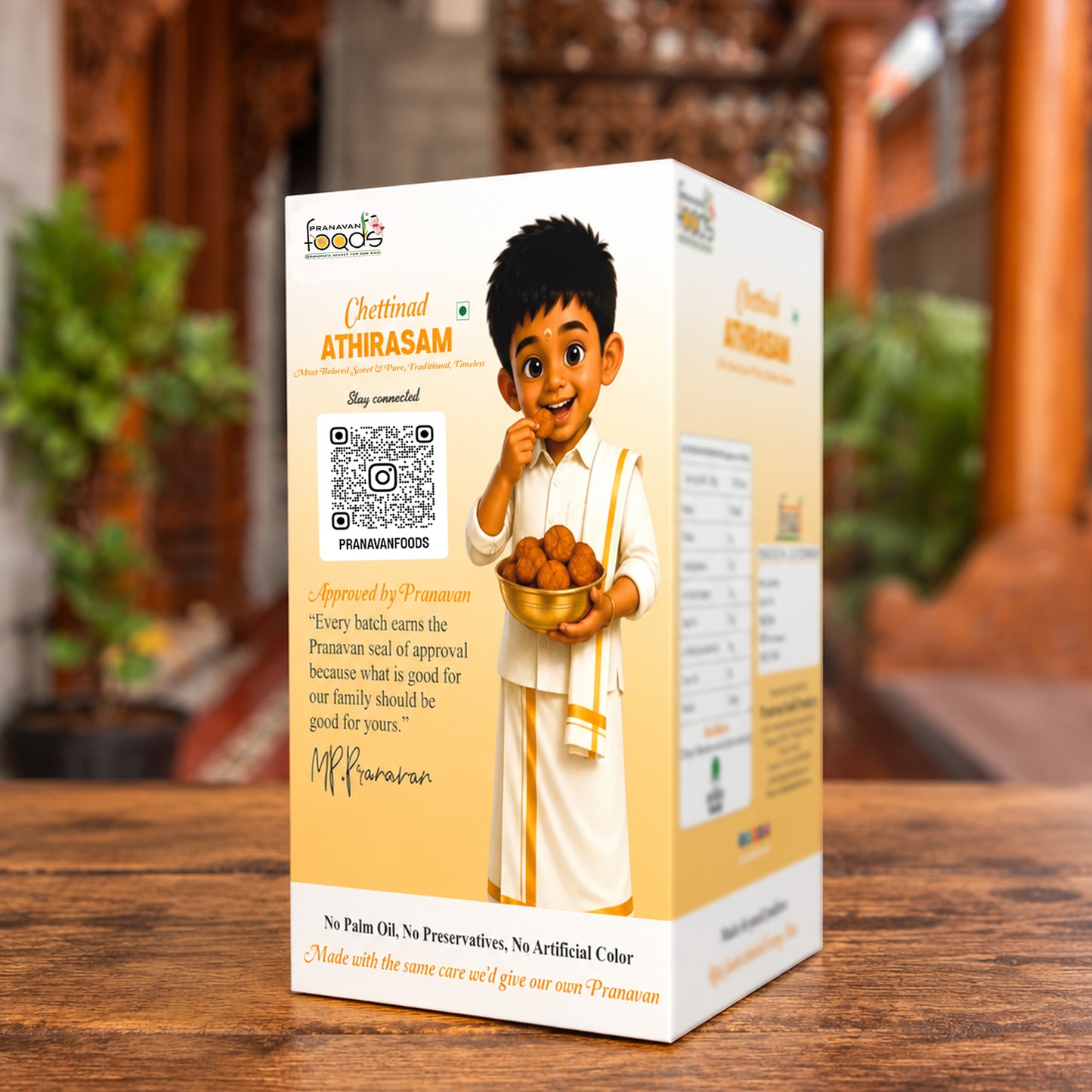 A boxed snack product labeled 'Chettinad Athirasam' stands on a wooden surface with a blurred outdoor background. The box is cream and white with orange text and features an illustrated boy holding a bowl of round sweets, plus a QR code and approval quote.