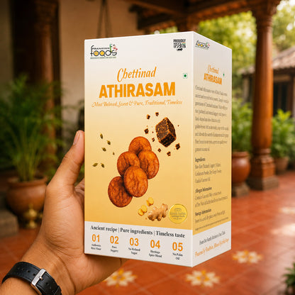 A hand holds a box of Chettinad Athirasm (traditional Indian fried snacks) against a blurred outdoor background with potted plants and a wooden structure.