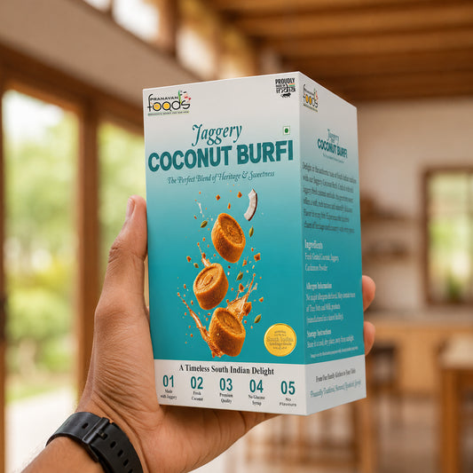 A hand holding a box of Coconut Burfi against a blurred outdoor background. The packaging is white and teal with illustrations of coconut burfi pieces and the brand name 'Pranavan Foods' visible at the top left.
