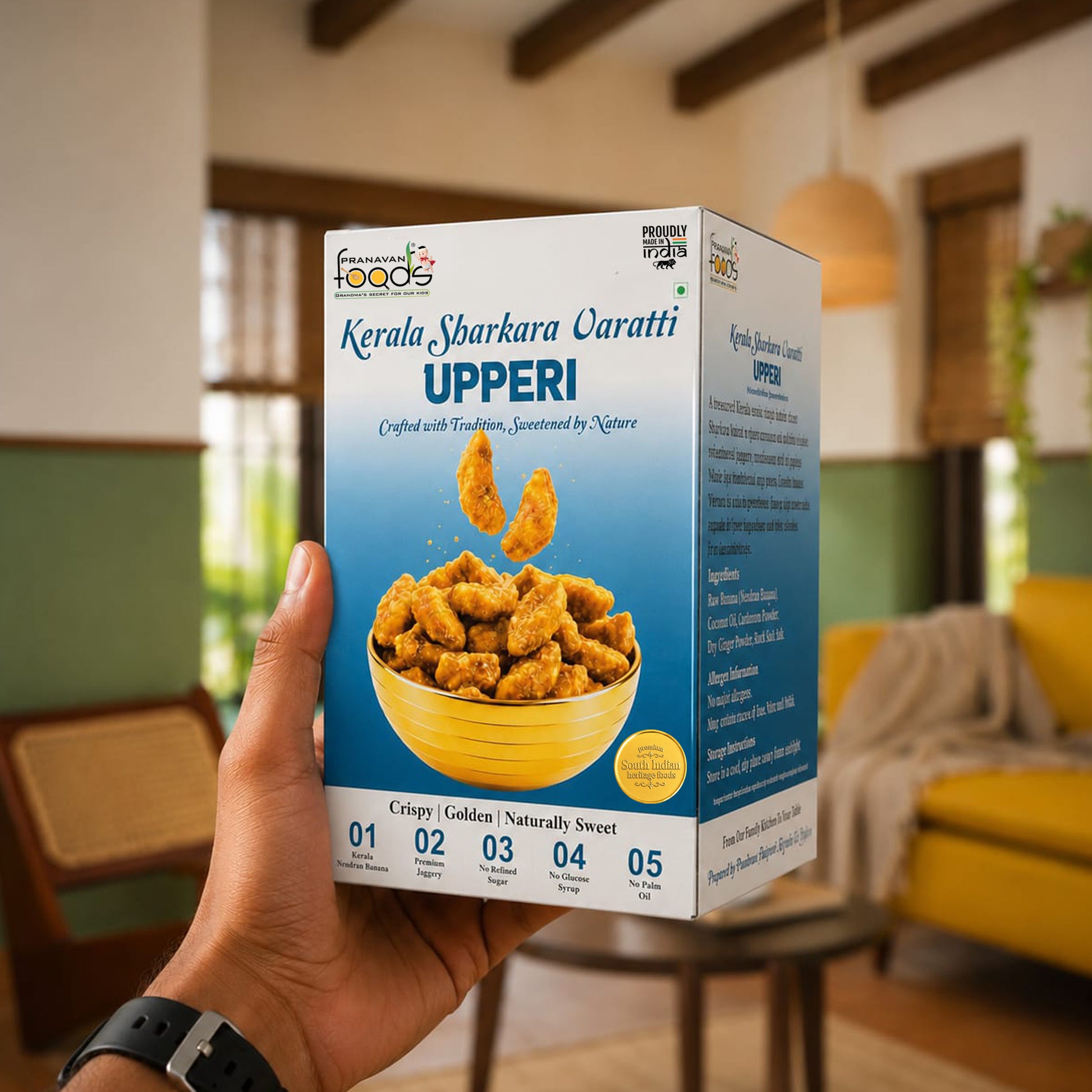 A hand holds a box of Kerala Sharkara varatti Upperi crispy fried snacks in a cozy, softly blurred indoor setting with warm lighting and wooden furniture.