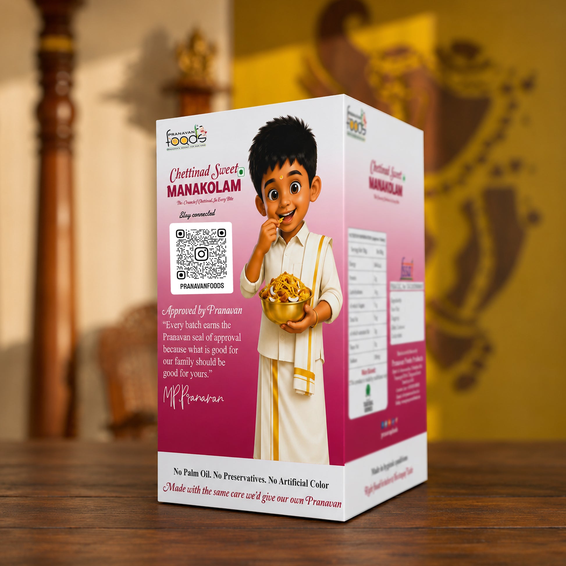 A boxed food product labeled 'Chettinad Sweet, MANAKOLAM' stands on a wooden surface. The package is predominantly pink and white with an illustrated boy holding a bowl of sweets, a QR code, and approval text.