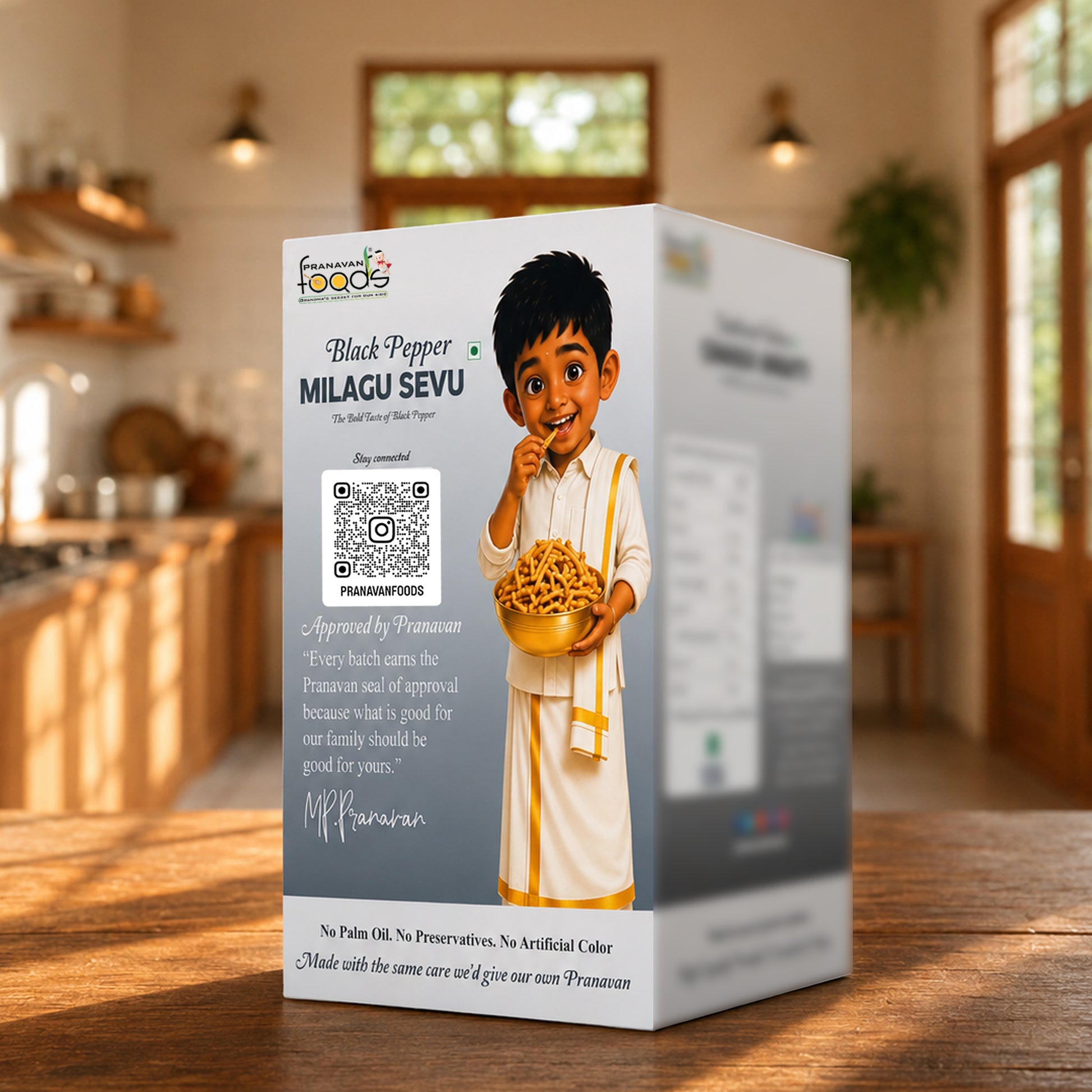 A boxed Pranavan food product labeled 'Black Pepper MILAGU SEVU' stands on a wooden table in a warm, blurred kitchen setting. The box is white with gray side panels, printed text, a QR code, and an illustration of a child holding a bowl of pepper sevu.