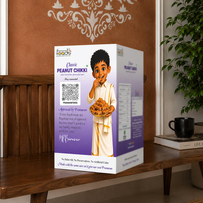 A box of Pranavan Classic Peanut Chikki stands on a wooden table in a home setting, with a brown wall, decorative wall paneling, a potted plant, and a black mug in the background.