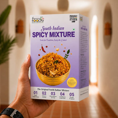 A hand holds a box of Freshly From The Farm South Indian Spicy Mixture against a blurred indoor background with warm lighting and arches.