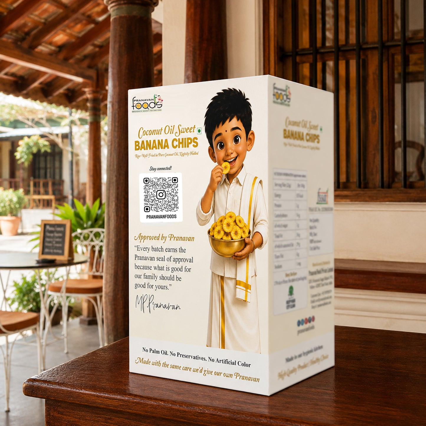 A white rectangular box of Pranavan Coconut Oil Sweet Banana Chips stands on a wooden table in a restaurant setting. The front of the box features a cartoon illustration of a boy holding a bowl of banana chips, along with product text and a QR code.