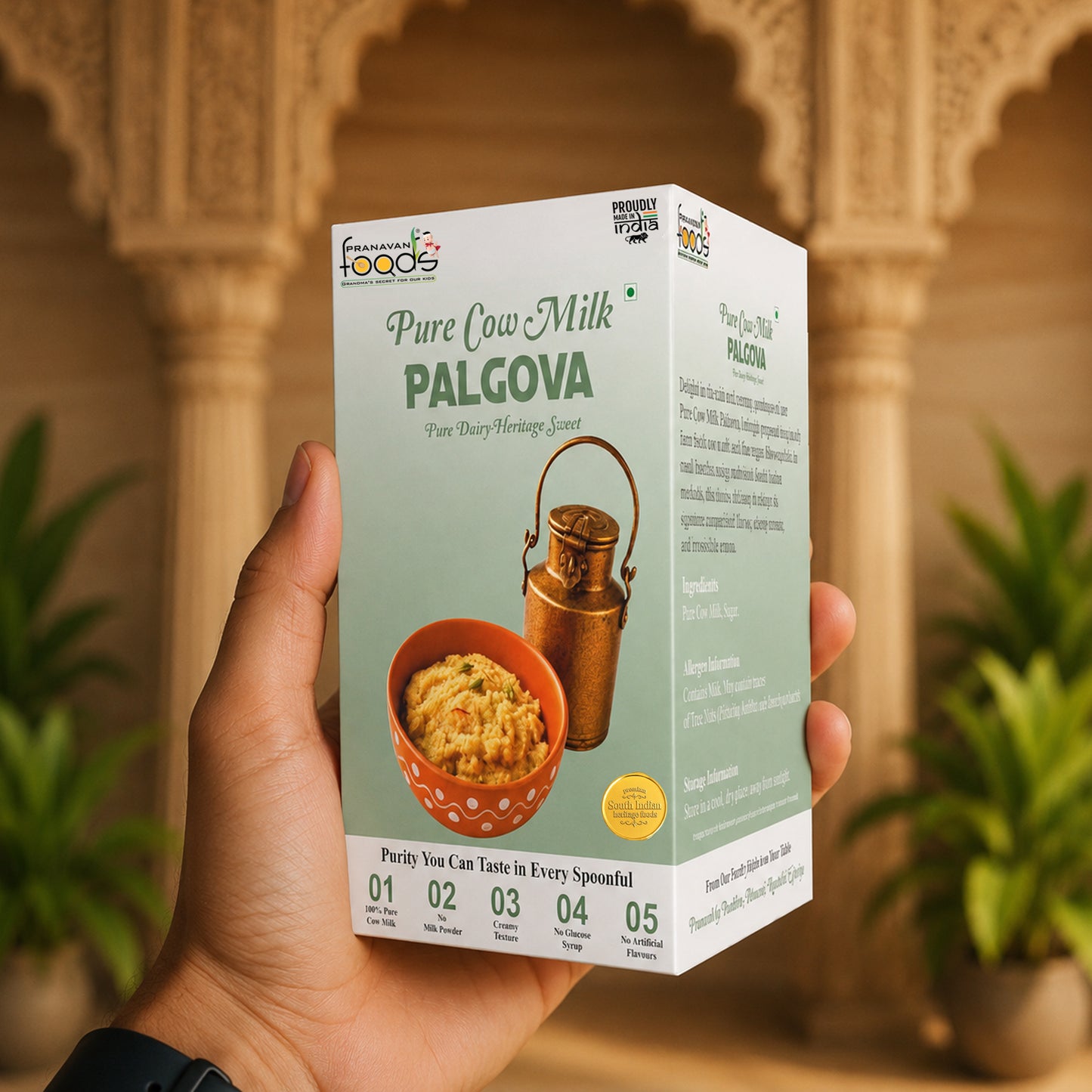 A hand holds a box of Pranavan Foods Pure Cow Milk Palkova (pure dairy) against a warm, blurred background of an Indian courtyard with arches and potted plants.