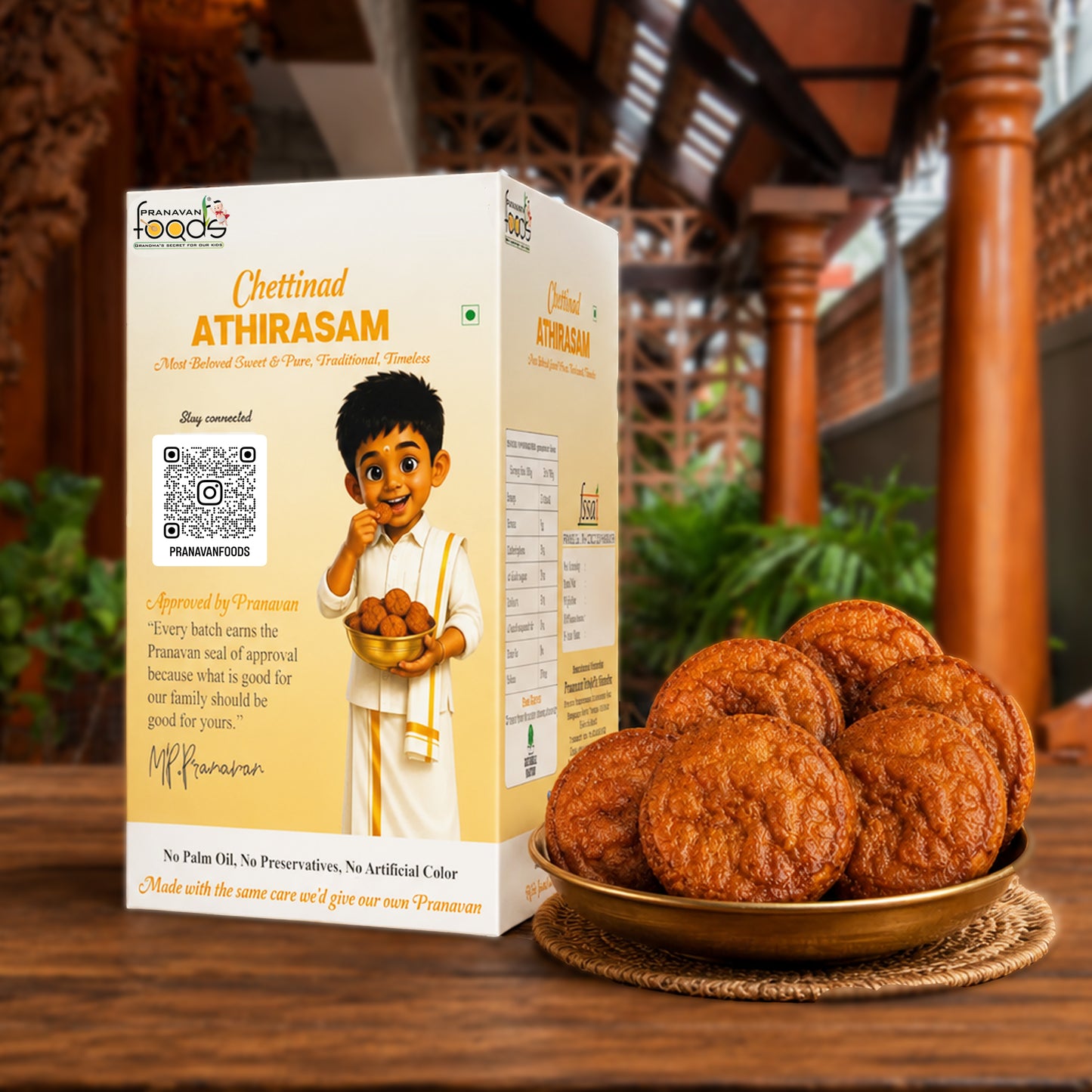 A box of Pranavan Foods Chettinad Athirasam ladoo cookies stands on a wooden table next to a gold plate filled with the round, golden-brown cookies. The background shows a blurred traditional wooden structure.
