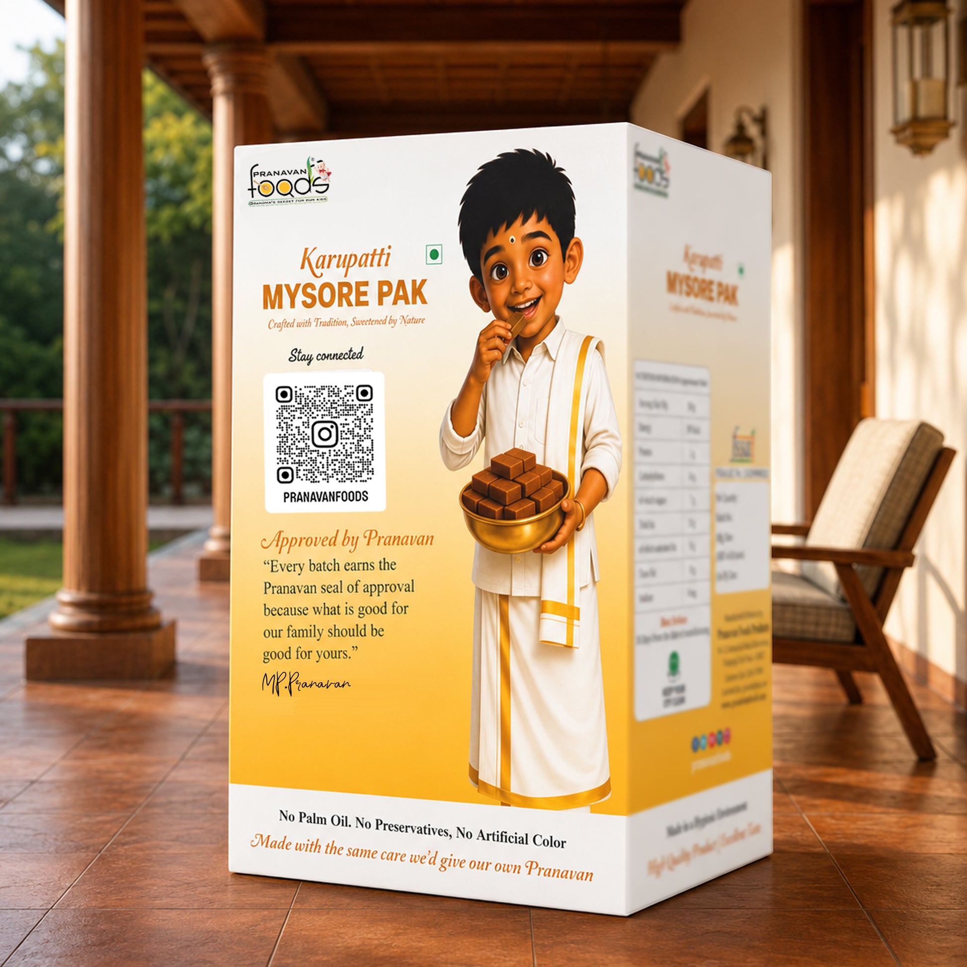 A boxed food product labeled 'Karupatti Mysore Pak' stands on a wooden porch. The package is predominantly white and warm yellow/gold, with printed illustrations of a child in white clothing holding a bowl of food, plus a QR code and text.