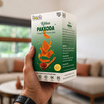 A hand holds a box of Pranavan Foods Ribbon Pakkoda in a brightly lit indoor setting. The packaging is green and white with illustrations of golden-brown pakkoda crackers and a small yellow seal.