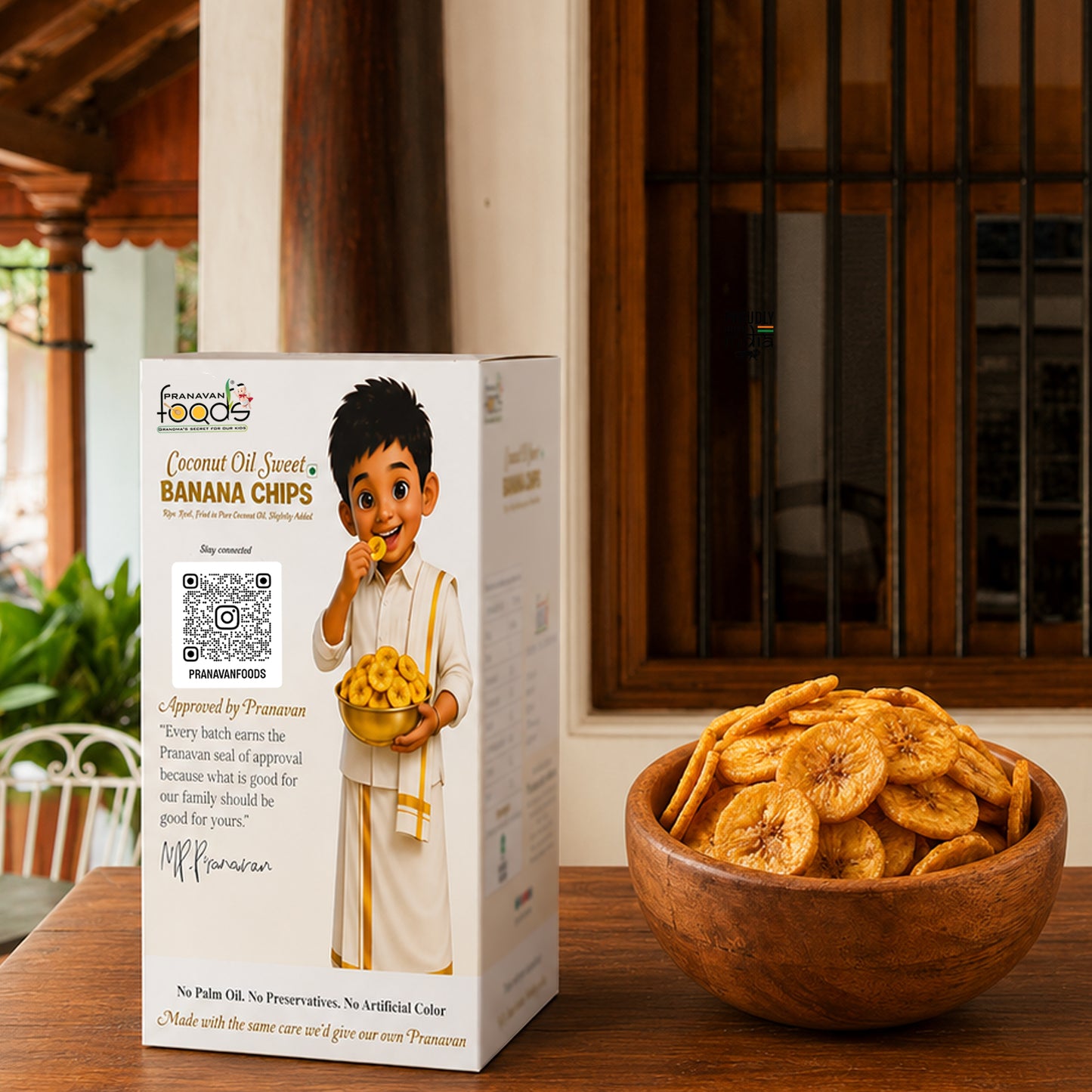 A box of Prancess food banana chips stands on a wooden table beside a wooden bowl filled with sliced banana chips. The box features a child character in traditional clothing and includes a QR code and approval quote from Prancess.