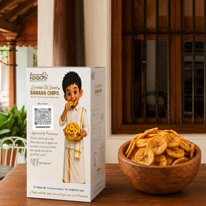 A box of Prancess food banana chips stands on a wooden table beside a wooden bowl filled with sliced banana chips. The box features a child character in traditional clothing and includes a QR code and approval quote from Prancess.